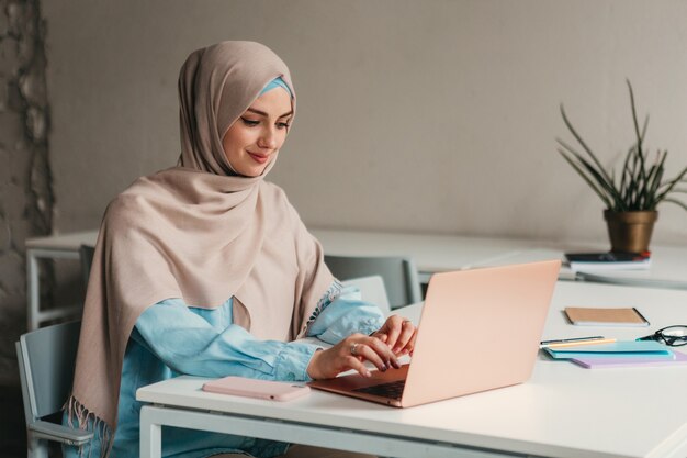 Young pretty modern muslim woman in hijab working on laptop in office room, education online