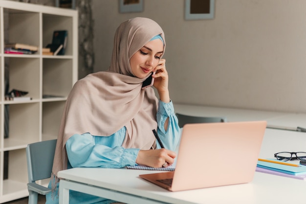 Young pretty modern muslim woman in hijab working on laptop in office room, education online