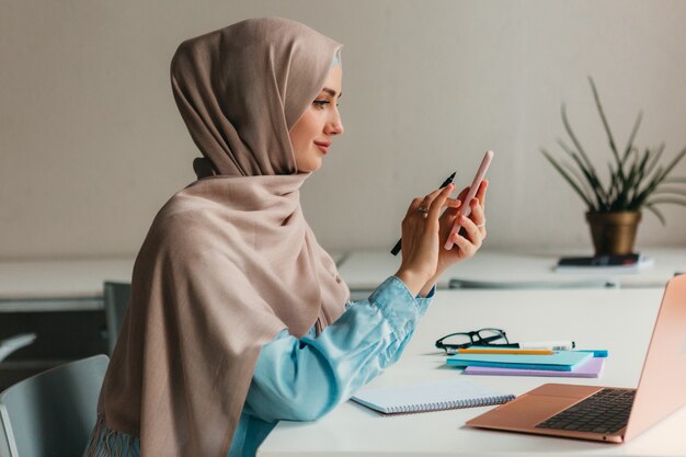 Young pretty modern muslim woman in hijab working on laptop in office room, education online