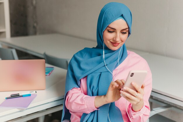 Young pretty modern muslim woman in hijab working on laptop in office room, education online