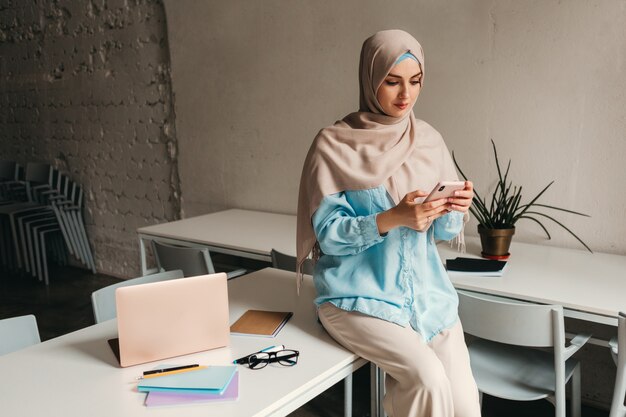 Young pretty modern muslim woman in hijab working in office room, education online