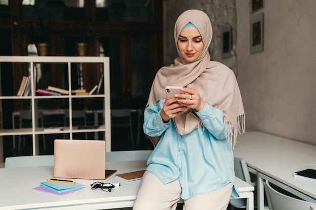 Young pretty modern muslim woman in hijab working in office room, education online