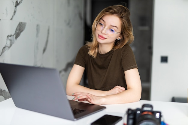 Young pretty photographer processing images on laptop, looking at photocamera at home