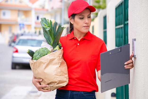 Young pretty postwoman holding paper bag and ringing doorbell. Confident brunette deliverywoman in red uniform doing her job and delivering order on foot. Food delivery service and post concept