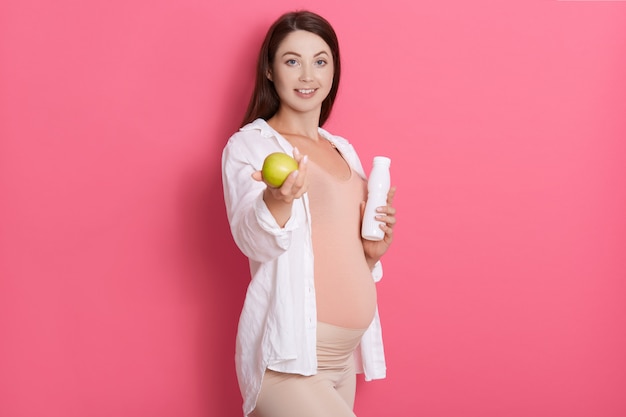 Young pretty pregnant woman holding green apple while posing isolated over pink