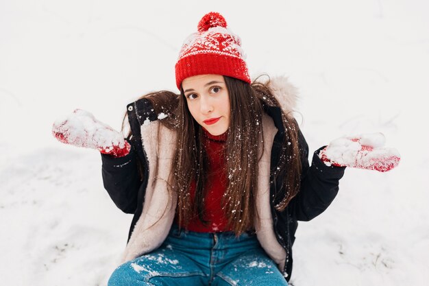 Young pretty puzzled woman in red mittens and knitted hat wearing winter coat sitting on snow in park, warm clothes