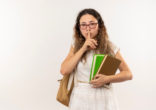 Young pretty schoolgirl wearing glasses and back bag holding books and gesturing silence isolated on white background with copy space