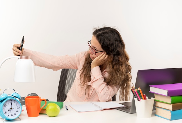 Giovane studentessa graziosa con gli occhiali seduto alla scrivania con strumenti di scuola facendo i compiti prendendo selfie isolati su sfondo bianco