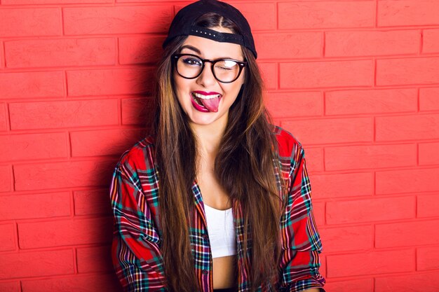 Young pretty seductive hipster woman having fun and showing long tongue and winked, wearing plaid shirt and clear glasses, long hairs and bright makeup.