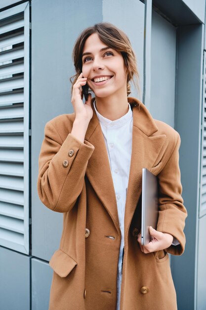 Young pretty smiling businesswoman in coat with laptop happily talking on cellphone outdoor