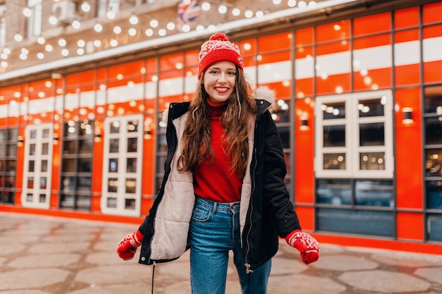 Молодая симпатичная улыбающаяся возбужденная счастливая женщина в красных рукавицах и вязаной шапке в зимнем пальто гуляет по городской рождественской улице, модная тенденция в стиле теплой одежды