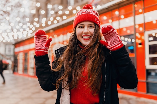 Молодая симпатичная улыбающаяся возбужденная счастливая женщина в красных рукавицах и вязаной шапке в зимнем пальто гуляет по городской рождественской улице, модная тенденция в стиле теплой одежды