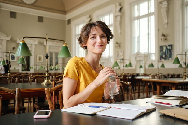 Молодая симпатичная улыбающаяся студентка с водой в руке радостно смотрит в камеру во время учебы в библиотеке университета