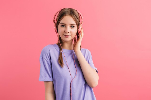 Young pretty smiling girl with two braids in lilac t shirt listening music in headphones happily looking in camera over pink background isolated