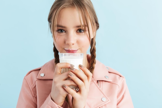 Young pretty smiling girl with two braids in pink leather jacket with milk mustache holding glass in hand while dreamily looking in camera over blue background