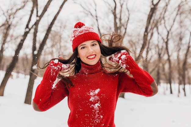 赤いミトンと帽子をかぶった若いかわいい笑顔の幸せな女性は、雪の中で公園を歩いて、暖かい服を着て、楽しんで、長い髪を振って、ニットのセーターを着ています