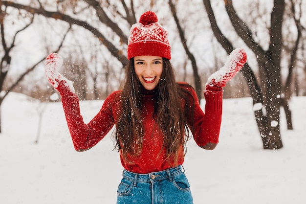 雪の中で公園を歩いて、暖かい服を着て、楽しんで、赤いミトンと帽子をかぶった若いかなり笑顔の幸せな女性