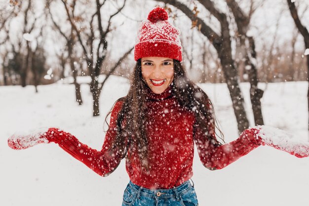 雪の中で公園を歩いて、暖かい服を着て、楽しんで、赤いミトンと帽子をかぶった若いかなり笑顔の幸せな女性