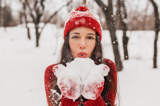 Young pretty smiling happy woman in red mittens and hat wearing knitted sweater walking in park in snow, warm clothes, having fun