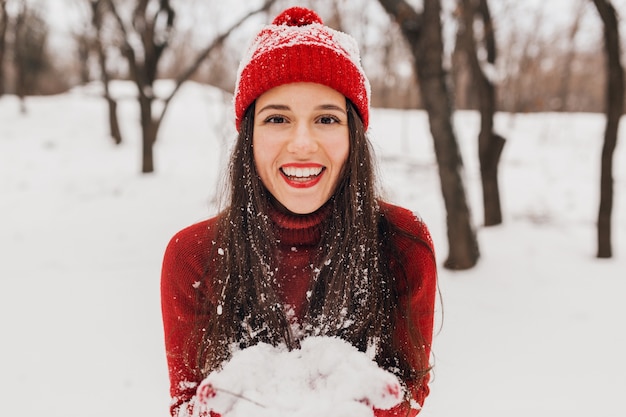 Young pretty smiling happy woman in red mittens and hat wearing knitted sweater walking in park in snow, warm clothes, having fun