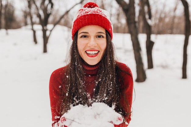 Young pretty smiling happy woman in red mittens and hat wearing knitted sweater walking in park in snow, warm clothes, having fun