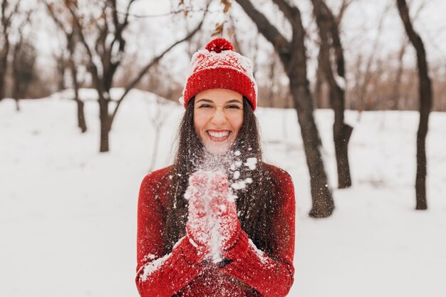 Young pretty smiling happy woman in red mittens and hat wearing knitted sweater walking in park in snow, warm clothes, having fun