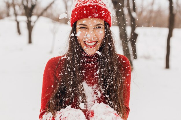 雪の中で公園を歩いて、暖かい服を着て、楽しんで、赤いミトンと帽子をかぶった若いかなり笑顔の幸せな女性