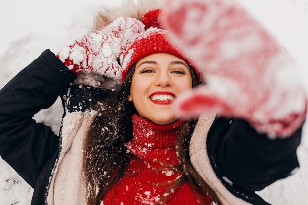 Young pretty smiling happy woman in red mittens and knitted hat wearing winter coat lying in park in snow, warm clothes, view from above