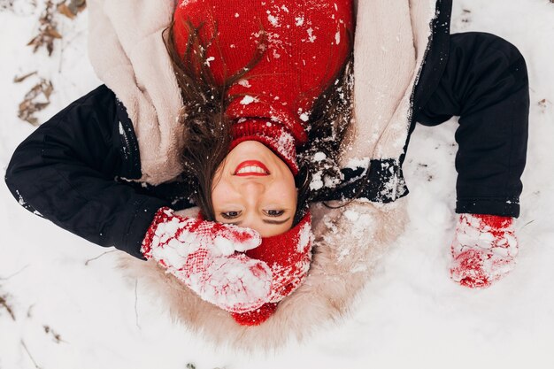 雪、暖かい服、上からの眺めの公園で横たわっている冬のコートを着て赤いミトンとニット帽の若いかなり笑顔の幸せな女性