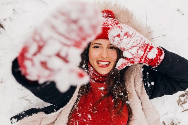 Young pretty smiling happy woman in red mittens and knitted hat wearing winter coat lying in park in snow, warm clothes, view from above