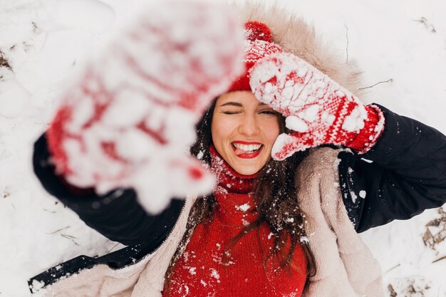 Young pretty smiling happy woman in red mittens and knitted hat wearing winter coat lying in park in snow, warm clothes, view from above