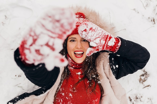 Молодая довольно улыбающаяся счастливая женщина в красных рукавицах и вязаной шапке в зимнем пальто, лежа в парке в снегу, теплой одежде, вид сверху