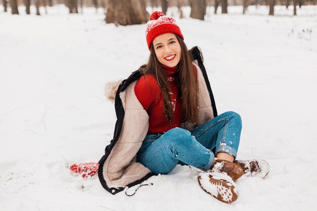 Young pretty smiling happy woman in red mittens and knitted hat wearing winter coat sitting on snow in park, warm clothes