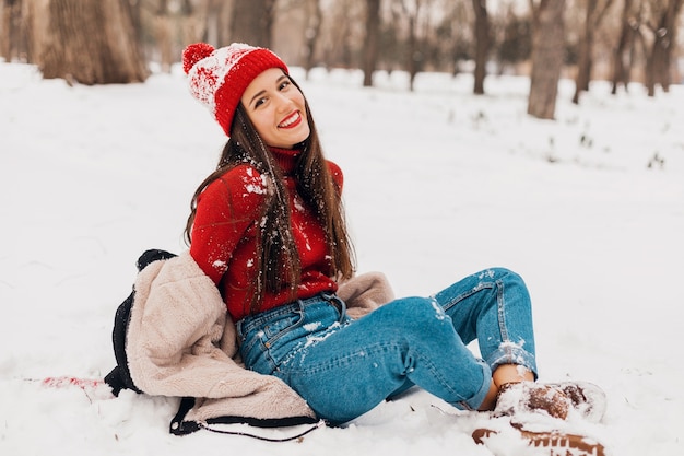 Foto gratuita giovane donna felice abbastanza sorridente in guanti rossi e cappello lavorato a maglia che indossa cappotto invernale seduto sulla neve nel parco, vestiti caldi