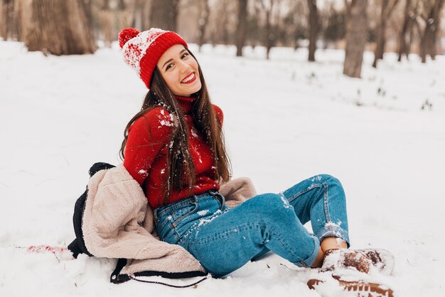 Young pretty smiling happy woman in red mittens and knitted hat wearing winter coat sitting on snow in park, warm clothes