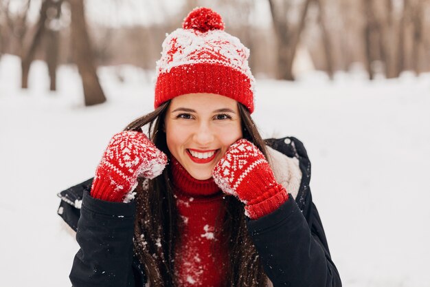 Young pretty smiling happy woman in red mittens and knitted hat wearing winter coat sitting on snow in park, warm clothes