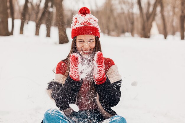 Young pretty smiling happy woman in red mittens and knitted hat wearing winter coat sitting on snow in park, warm clothes