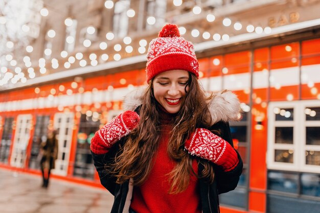 Young pretty smiling happy woman in red mittens and knitted hat wearing winter coat walking in city Christmas street, warm clothes style fashion trend