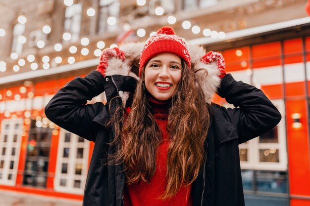 Young pretty smiling happy woman in red mittens and knitted hat wearing winter coat walking in city Christmas street, warm clothes style fashion trend