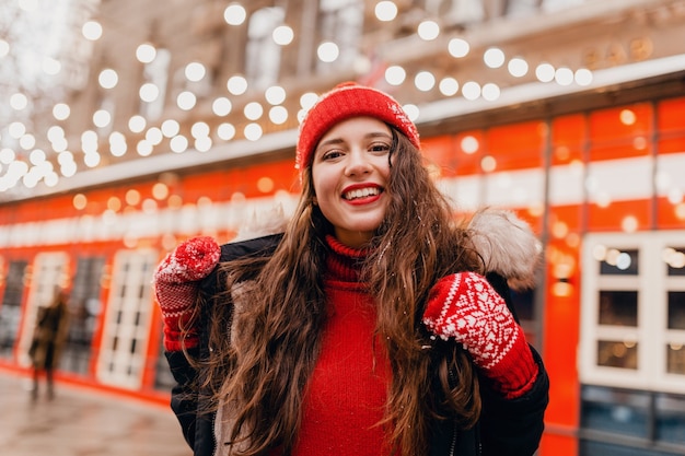 Молодая красивая улыбающаяся счастливая женщина в красных рукавицах и вязаной шапке в зимнем пальто гуляет по городской улице, теплая одежда