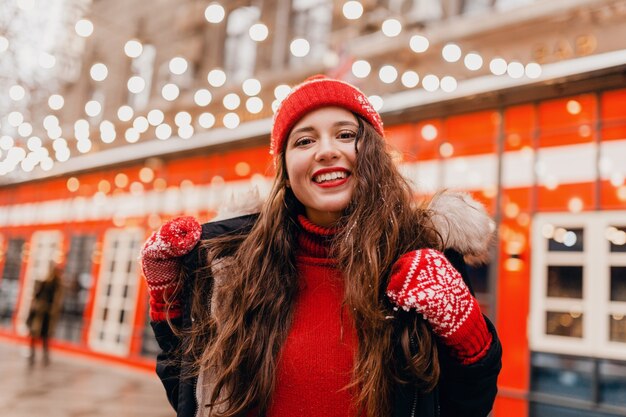 赤いミトンとニット帽を身に着けている若いかわいい笑顔の幸せな女性は、街の通りを歩いて、暖かい服を着て冬のコートを着ています