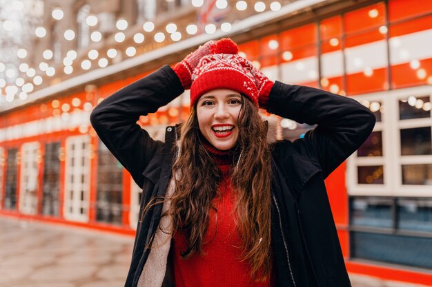 Молодая красивая улыбающаяся счастливая женщина в красных рукавицах и вязаной шапке в зимнем пальто гуляет по городской улице, теплая одежда