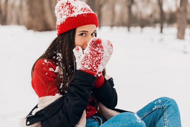 赤いミトンと冬のコートを着て、公園を歩いて、暖かい服を着て雪で遊んで、手を暖めるニット帽の若いかなり笑顔の幸せな女性