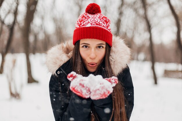 Молодая довольно улыбающаяся счастливая женщина в красных рукавицах и вязаной шапке в зимнем пальто, гуляет в парке, играет со снегом в теплой одежде