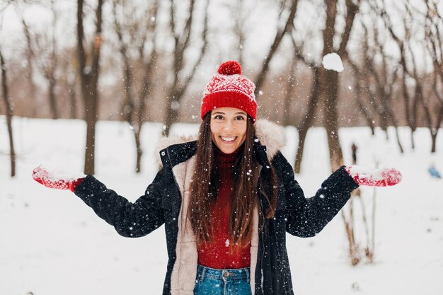 Young pretty smiling happy woman in red mittens and knitted hat wearing winter coat, walking in park, playing with snow in warm clothes