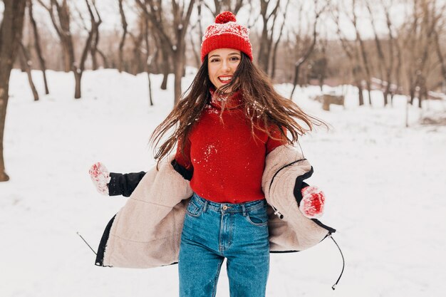 Young pretty smiling happy woman in red mittens and knitted hat wearing winter coat walking in park in snow, warm clothes, having fun