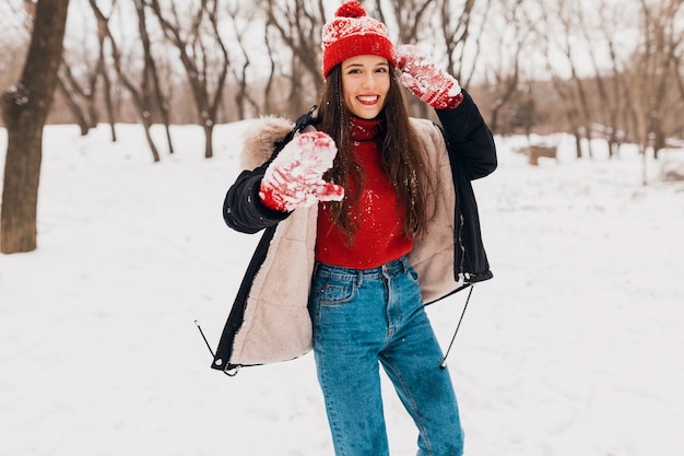 Young pretty smiling happy woman in red mittens and knitted hat wearing winter coat walking in park in snow, warm clothes, having fun