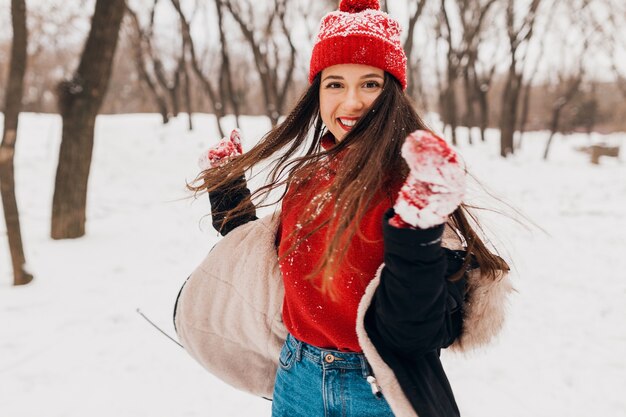 雪の中で公園を歩いて、暖かい服を着て、楽しんで、赤いミトンと冬のコートを着たニット帽の若いかわいい笑顔の幸せな女性