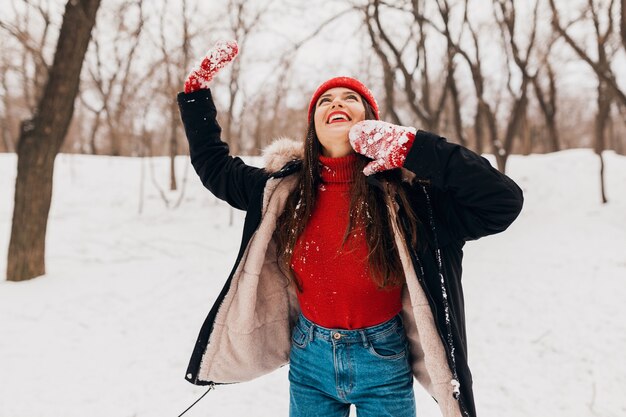Young pretty smiling happy woman in red mittens and knitted hat wearing winter coat walking in park in snow, warm clothes, having fun