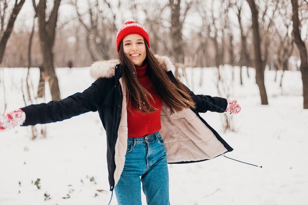 Молодая красивая улыбающаяся счастливая женщина в красных рукавицах и вязаной шапке в зимнем пальто, гуляет в парке в снегу, теплой одежде